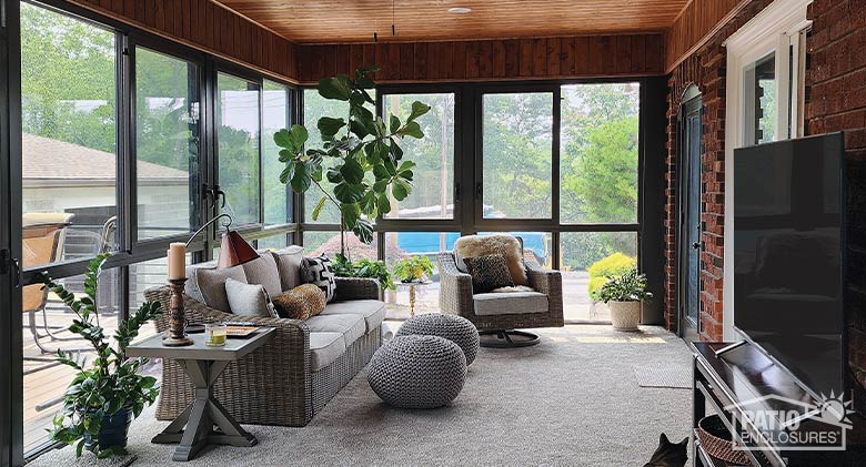 A comfortable living area with sofa, chair, ottomans, plants and TV in a four-season bronze sunroom with wood ceiling.