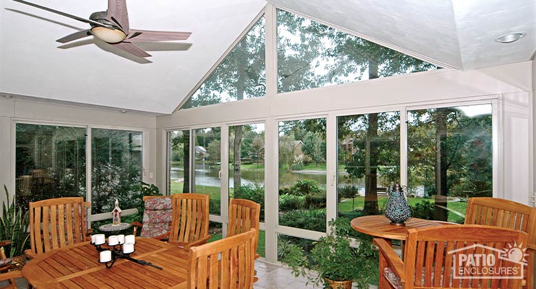 Comfortable furniture and plants in a second-story glass sunroom with an expansive view of trees and water beyond.