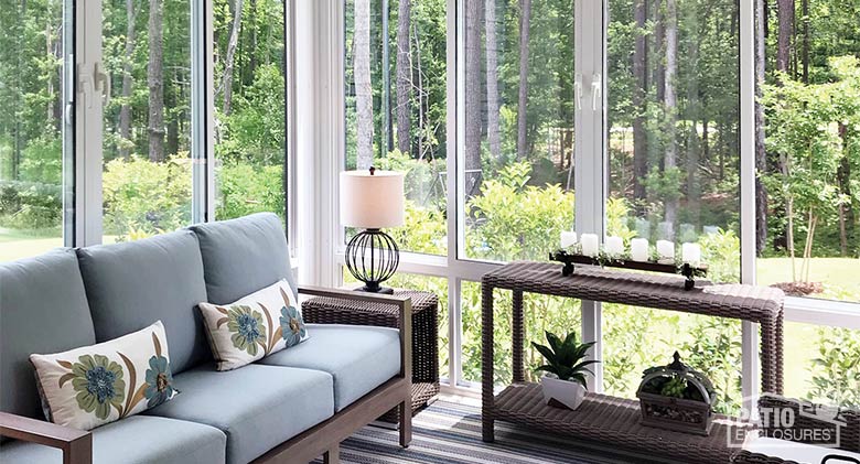 A light blue sofa with flower print pillows and two wicker side tables in a white glass three-season sunroom..