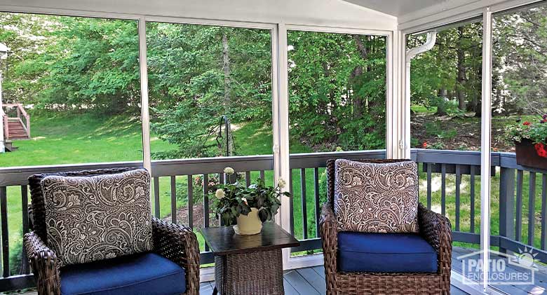 Two brown wicker chairs with blue cushions and neutral print cushions on the back inside a white screen room on a deck.