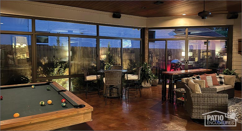 The interior of a sunroom at night furnished with a pool table, table and chairs and a comfortable seating area.