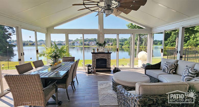  Interior of a glass sunroom with vaulted ceiling, ceiling fan, seating & dining areas, fireplace, and view of a large lake.