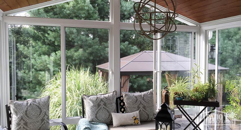 Interior of a three-season glass sunroom with wood ceiling, modern light fixture, loveseat, chair, and plants on a table..