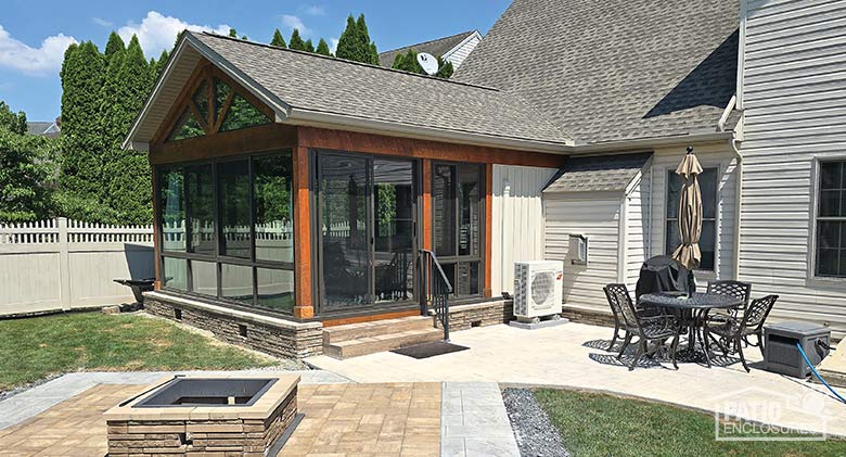 Glass three-season sunroom with wood trim, shingled, gable roof with glass wings, glass knee wall, and firepit/patio outside..