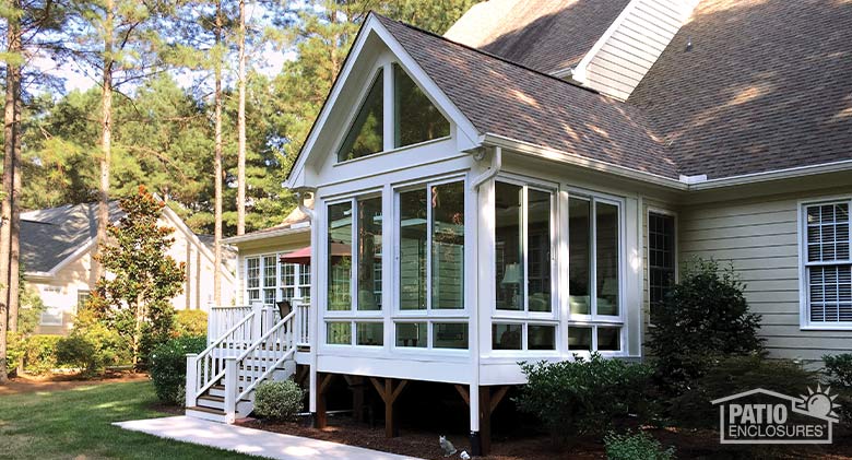 Tall white glass four-season room with vaulted, gable roof, glass wings, glass knee wall on beige home with deck.