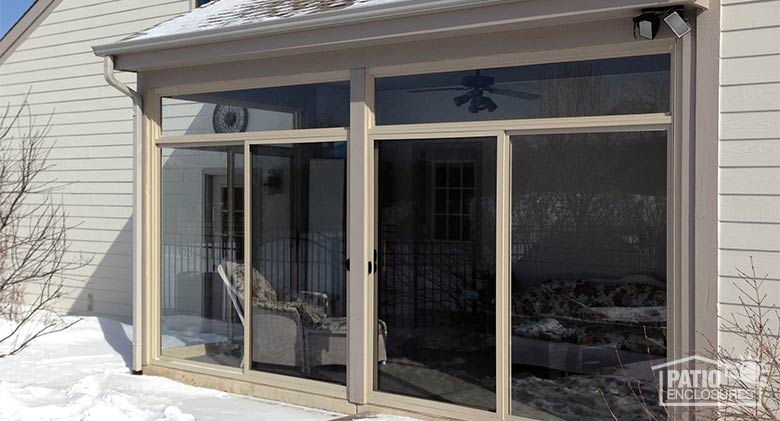 A glass patio enclosure with a beige frame surrounded by snow on the ground and roof.
