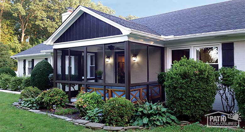 A screen room with gable roof and bronze frame surrounded by lush plantings and bushes.