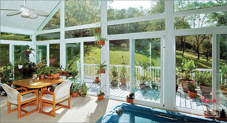 Ceiling fan above a table and chairs, lots of plants and a hot tub in the corner of a white sunroom with gable roof.