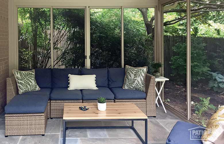 Shady interior of a screen room surrounded by trees and bushes, blue couch and wood coffee table in the room, tile floor.