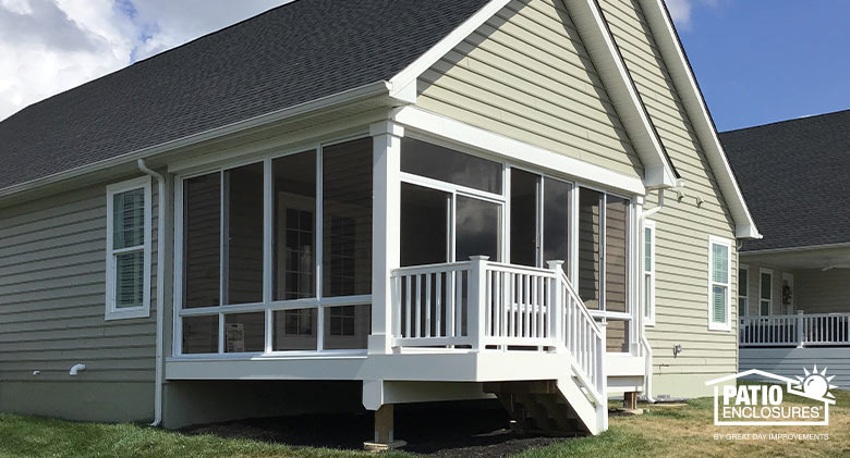 A few stairs lead down from a corner porch enclosed in glass on a tan home with gable roofs.