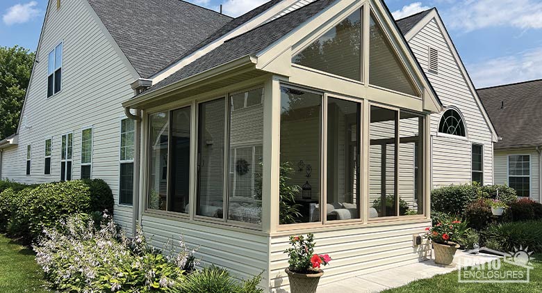 A sandstone three-season room built on an open patio with shingled gable roof and glass wings, potted flowers in front.
