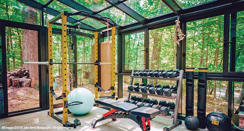 A brown solarium with glass roof furnished as a home gym with workout equipment and a view of a wooded yard on all sides.

