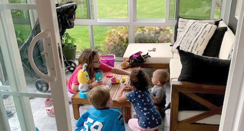 Four young children play with colorful toys on a coffee table in front of a sofa in a sunroom with glass walls.