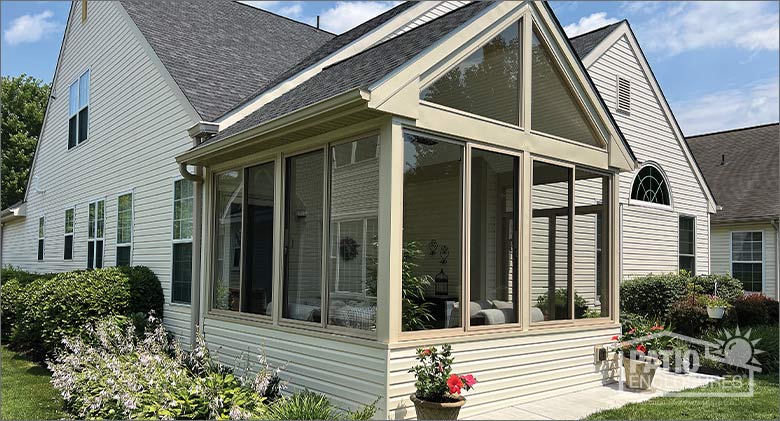 A tan house with a matching glass three-season porch with a gable roof, surrounded by neat landscaping.