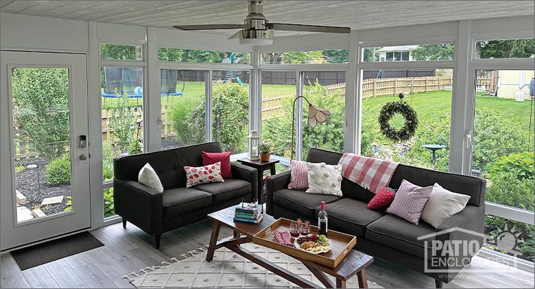 A glass sunroom with a brown sofa and loveseat with white and red pillows, and a coffee table with snacks and books.