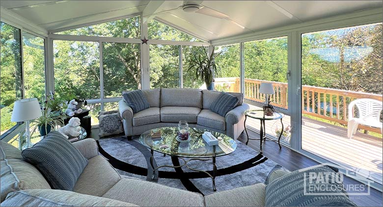 Comfortable curved couches in a glass sunroom with gable roof and a sweeping view of the deck and wooded backyard.