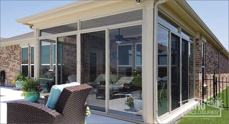 A glass three-season porch on a brick home, surrounded by a sunny open patio with comfortable chairs and plants.