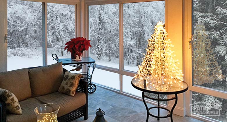 The interior of a sunroom with a poinsettia and a small lighted Christmas tree on a table; snow covered trees outside.