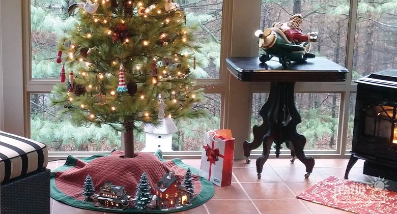 The bottom portion of a decorated Christmas tree with lighted ceramic houses, a fireplace, and other holiday décor nearby.