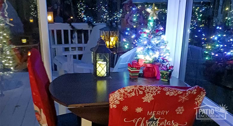 A small lighted Christmas tree, packages, and a candle on a tabletop; other holiday lights reflected in the windows.