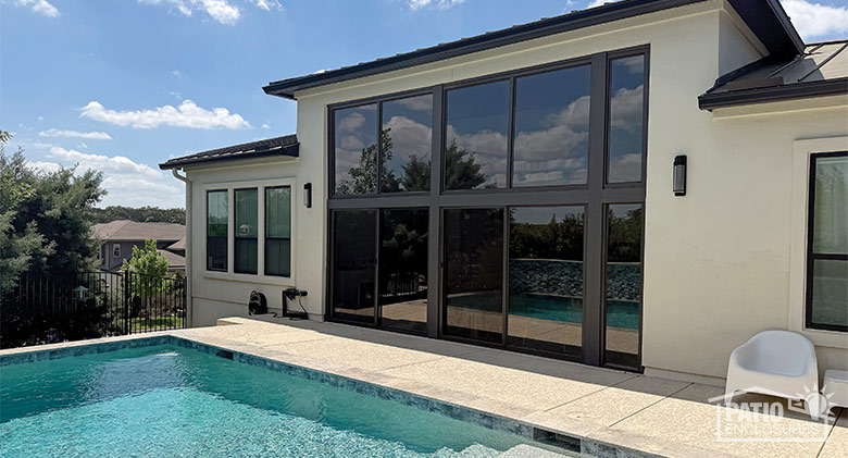 A large glass patio enclo sure with brown frame and an inground pool and patio in front of the room.