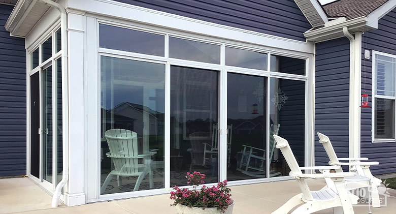 :A corner patio enclosed with a glass three-season room with white frame. A potted plant and two chairs outside on patio.