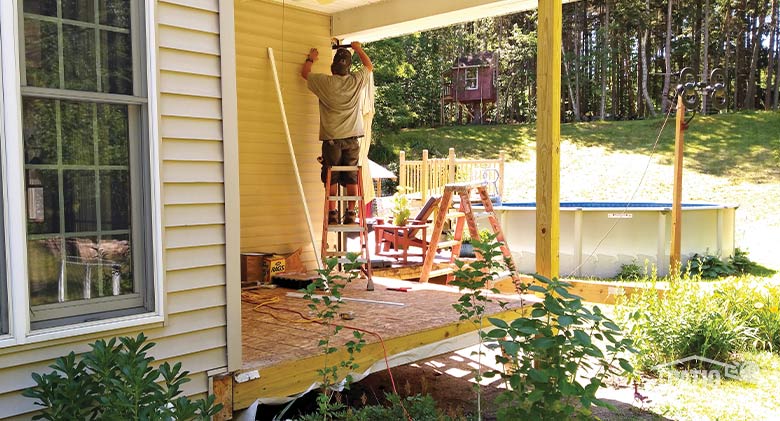 :A sunroom being installed on a corner porch of a house. The decking and suppport posts look new. Pool in background.