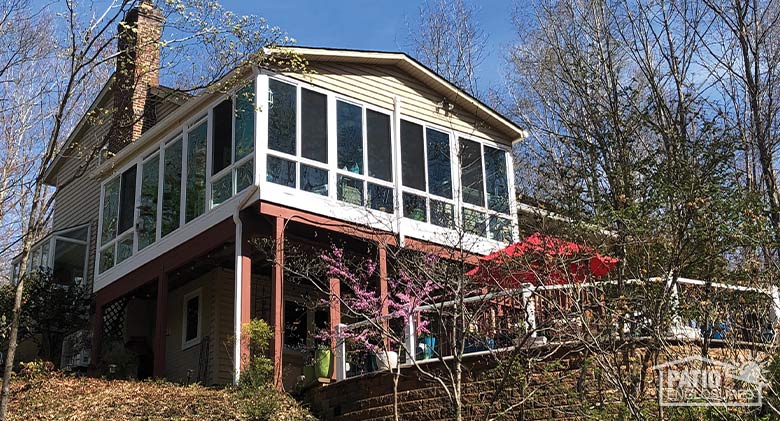 Large glass sunroom on second floor of a home on a hill, patio with table and red umbrella below and many trees.