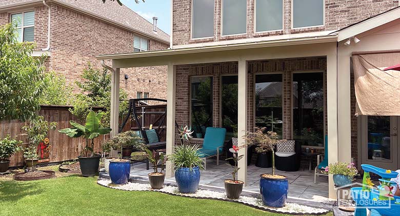 : A tan stationary patio cover on a brick home with potted plants in front and chairs with side tables under the cover.