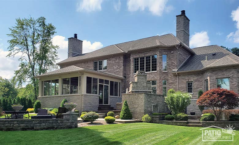 A large brick home on a sunny day with a raised patio enclosure, sweeping backyard, raised open patio, and formal plantings.