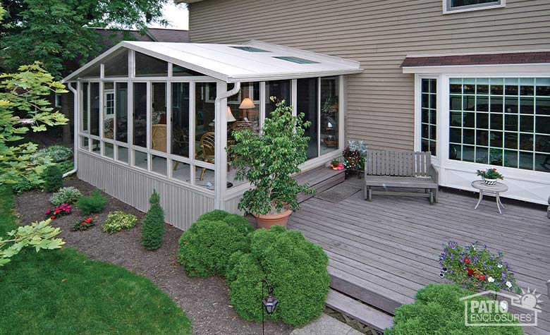 Looking down on a large beige sunroom with glass roof panels with mulched curved garden in front and deck on the right.