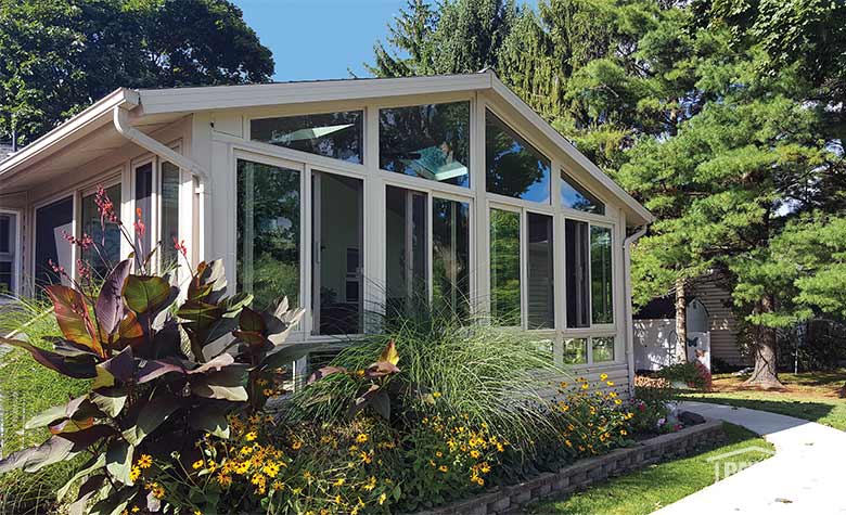 Yellow flowers, grasses, and a red-leafed tropical plants in front of a large beige sunroom with gable roof.