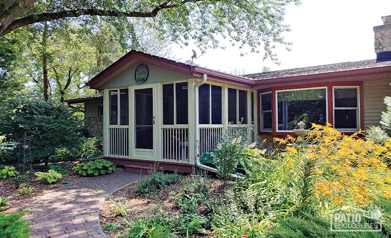 A garden of grasses, day lilies, and hostas with brick path in front of a screen room with gable roof and picket railing