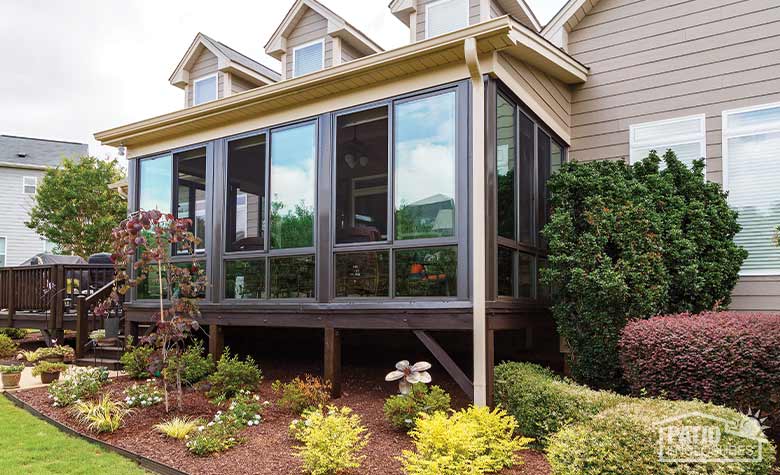 A mulched garden of small shrubs and trees in various colors in front of a raised brown glass porch enclosure.