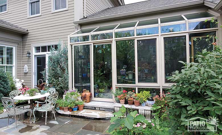 Potted plants, lush plantings, and a table and chairs on a patio in front of a glass bump-out solarium with beige frame.