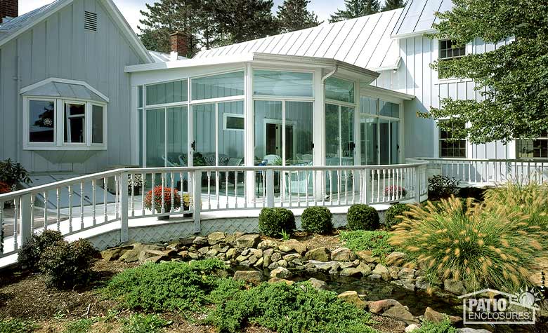 An unusually shaped glass sunroom surrounded by a large deck and a garden of shrubs, grasses, rocks, and small stream.