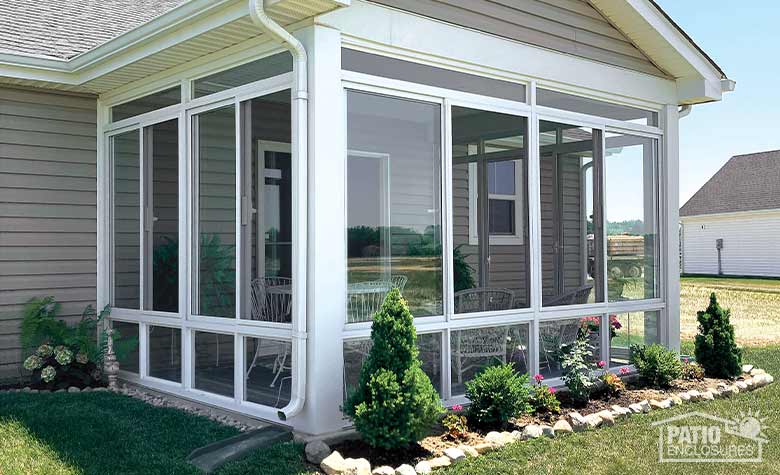 : A small flower bed with shrubs and annuals lined with rocks in front of a white glass sunroom with glass knee wall.