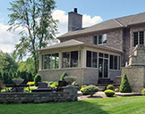 A large brick home on a sunny day with a raised patio enclosure, sweeping backyard, raised open patio, and formal plantings.