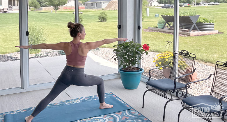 A woman in workout clothes striking a yoga pose in a sunroom with chairs on the side and a view of the backyard.