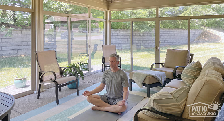 A middle-aged man sitting in lotus position on the floor of a sunroom meditating.
