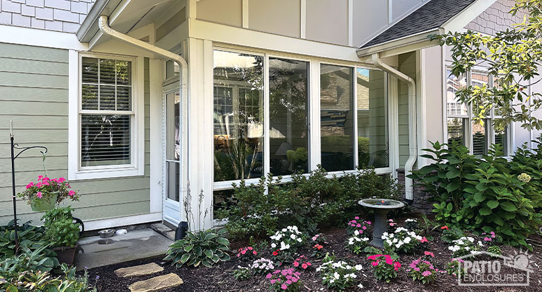 A white two-walled patio enclosure with a storm door on the left side and white and pink flowers in front.