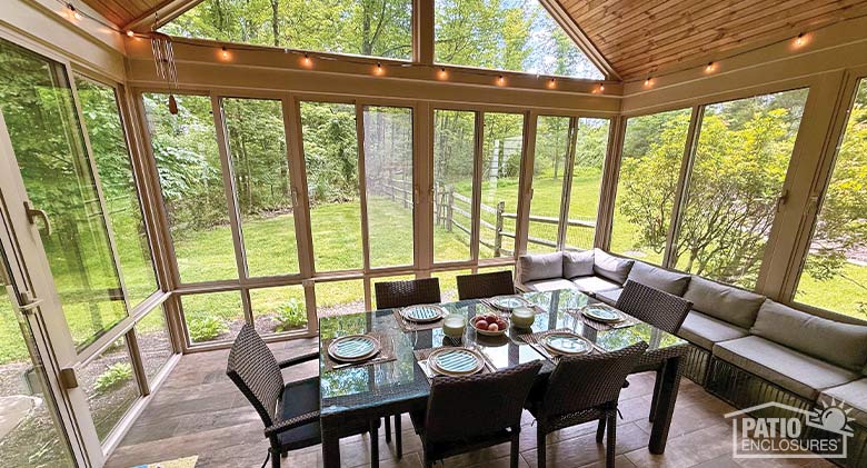 The interio a glass sunroom with beige frame and furnished with a long sofa and a dining table and chairs in the center.

