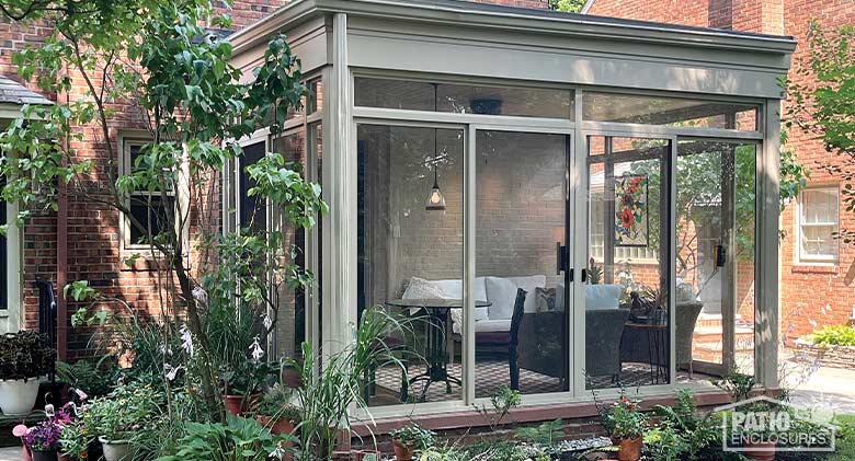 A tan glass sunroom patio enclosure surrounded by potted plants and small trees. Inside, a comfortable seating area.