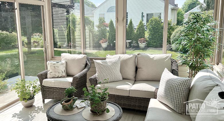 Comfortable chairs and loveseats with beige cushions inside a beige glass sunroom, coffee table with plants.