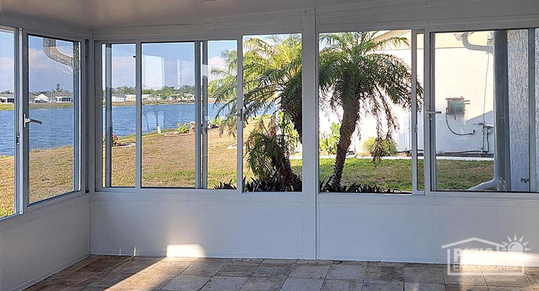 The interior of a white glass sunroom enclosure with solid knee wall. View through the windows of palm trees and lake.