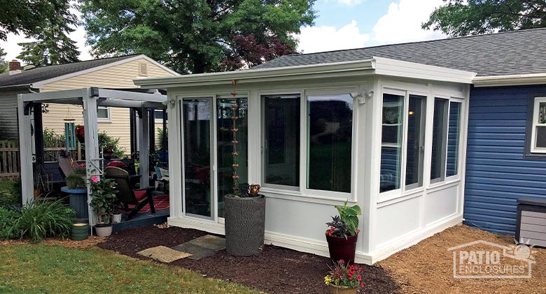 A white sunroom with shed roof and solid knee walls attached to a blue house and surrounded by neat plantings.