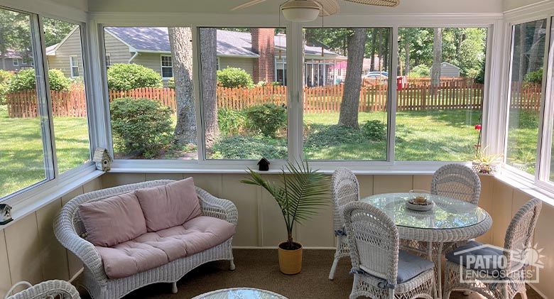 A three-season glass enclosure furnished with white wicker furniture and a plant.