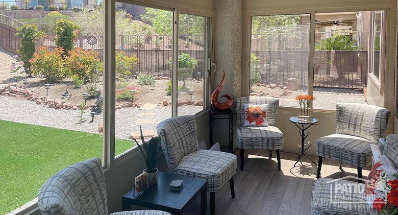 Views of a sunny landscape from a glass three-season room with solid knee wall and furnished with several chairs.