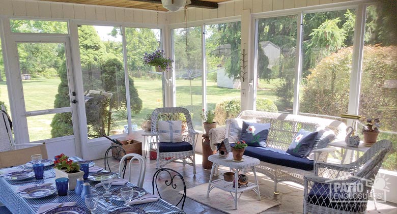 : A three-season porch enclosure furnished with white wicker furniture and blue cushions. A dining table in the foreground.