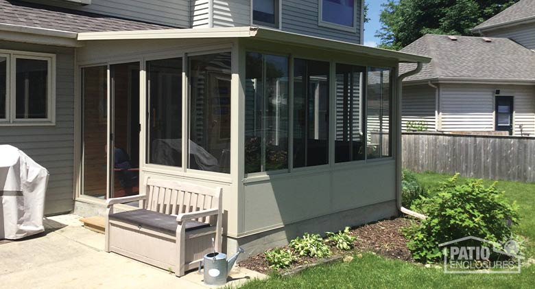 A tan sunroom with single-sloped roof and solid knee wall surrounded by neat flower beds, patio, and garden bench.